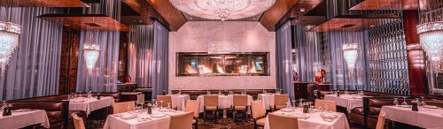 Upscale restaurant interior with white tablecloths, leather chairs, crystal chandeliers, a modern fireplace, and elegant sheer curtains.