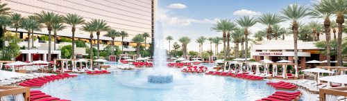 Luxury resort pool with a central fountain, palm trees, cabanas, and red lounge chairs.