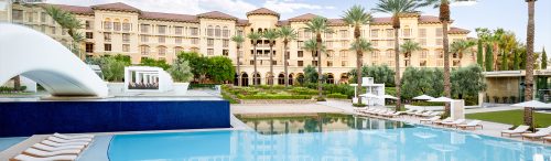 Luxury resort with a large pool, water slide, palm trees, and lounge chairs in front of an elegant Mediterranean-style building.