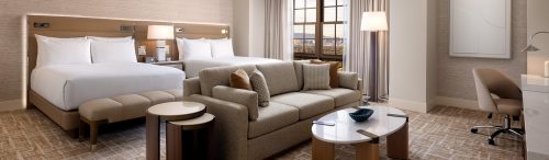 Modern hotel room with two beds, beige sofa, nesting tables, white marble coffee table, floor lamp, and large window with city view.