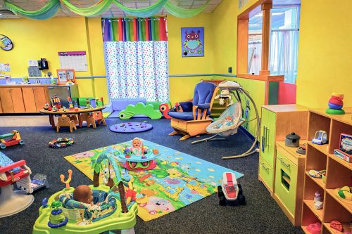 Bright daycare with yellow walls, colorful toys, and playful decor. Children sit in activity seats on a vibrant mat, creating a joyful atmosphere.