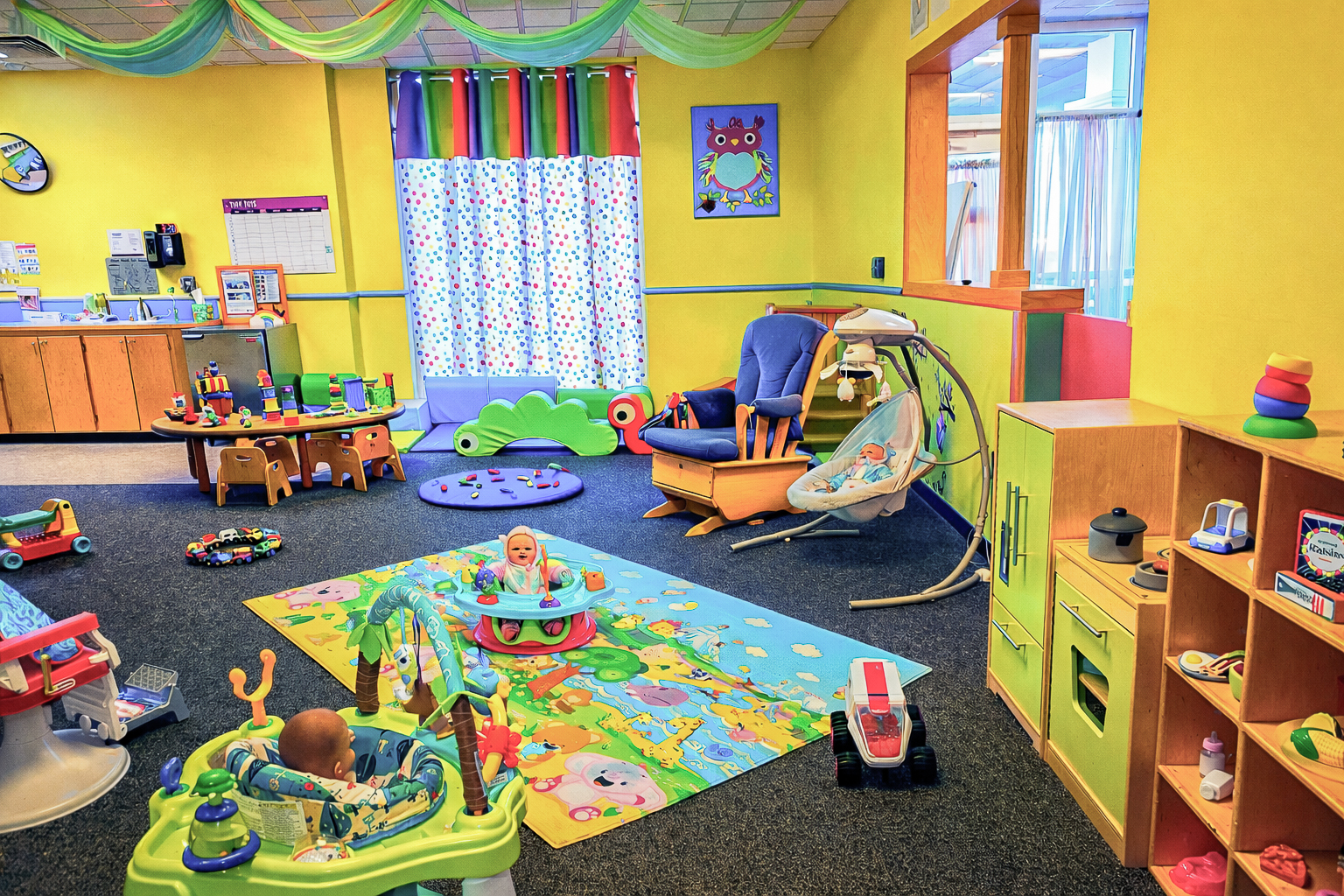 Bright daycare with yellow walls, colorful toys, and playful decor. Children sit in activity seats on a vibrant mat, creating a joyful atmosphere.