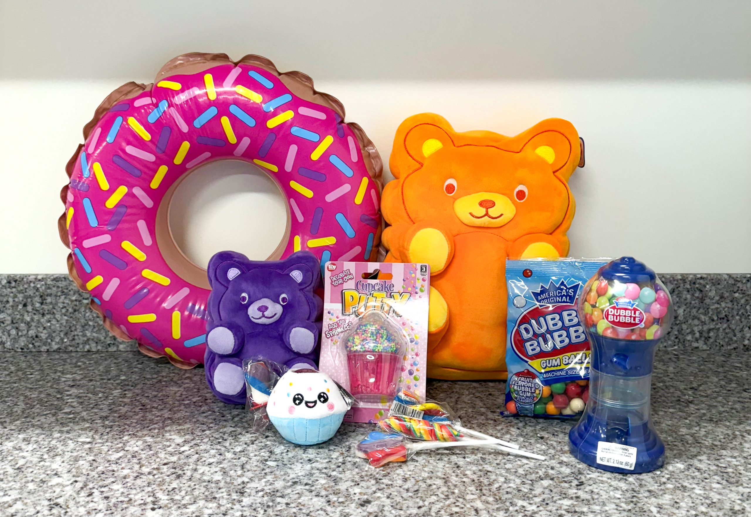 A colorful display of toys and candy on a countertop. Includes a pink donut float, bear cushions, plushies, rainbow putty, gum balls, and lollipops. Playful and fun.
