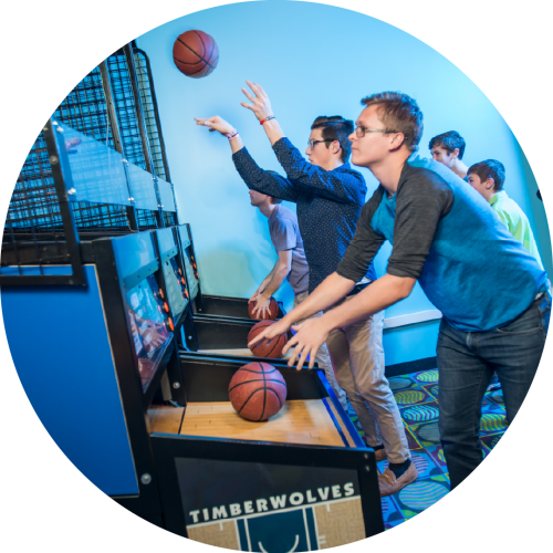 A group of young adults play arcade basketball, aiming energetically at the hoop. The lively scene has bright lighting and a fun, competitive atmosphere.