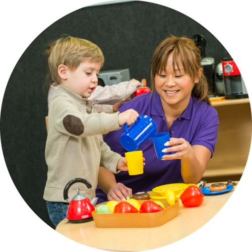 A woman and a child enjoy pretend play, pouring tea from a blue jug into cups. A tray of toy food creates a fun, playful atmosphere.