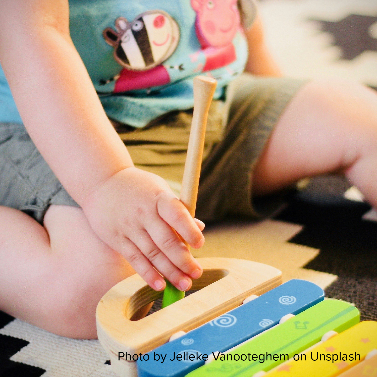 A child in a blue shirt plays with a colorful wooden toy xylophone, holding a mallet. The atmosphere is playful and creative.