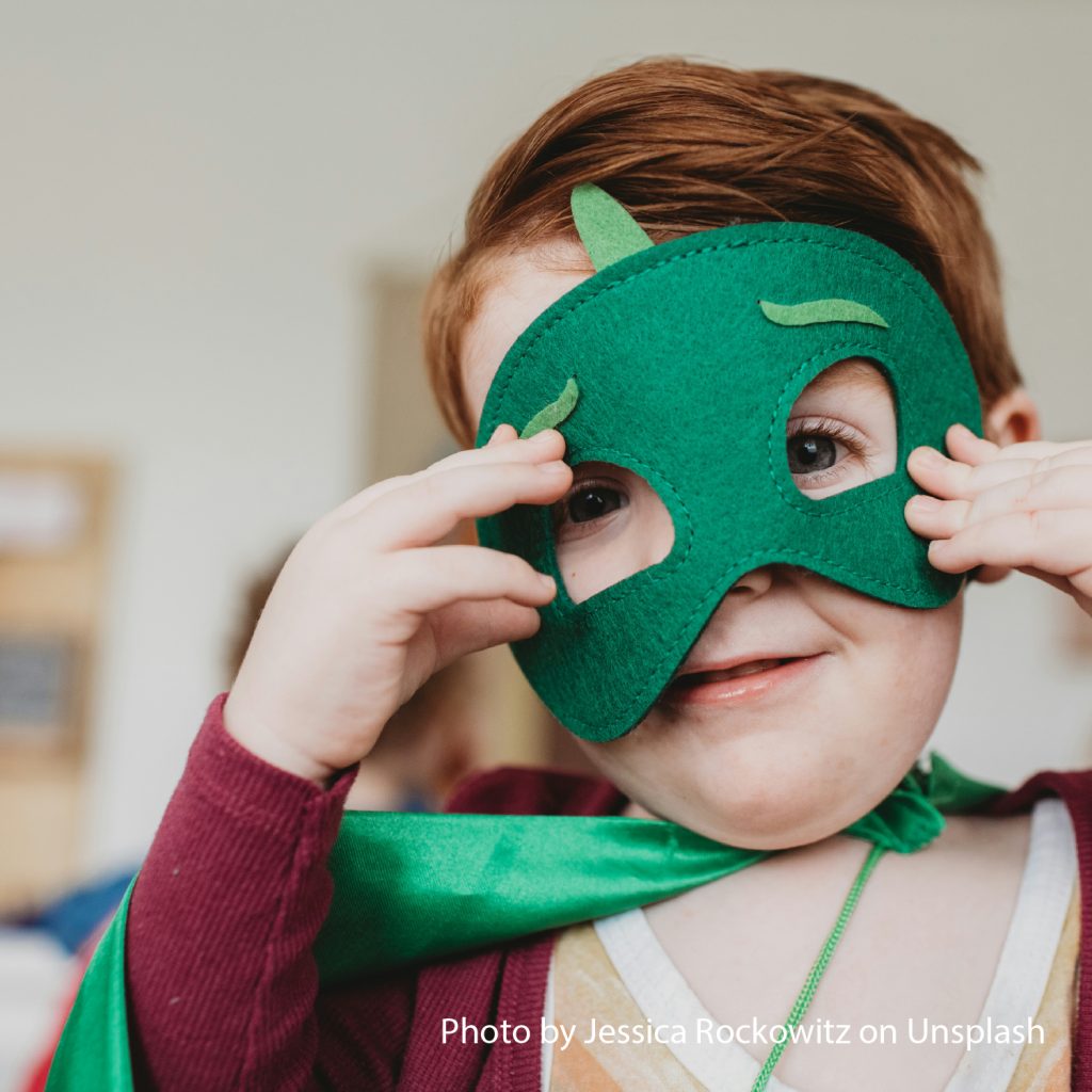 A child playfully wears a green superhero mask and cape, smiling with excitement. The setting is indoors, with a blurred background, creating a whimsical mood.