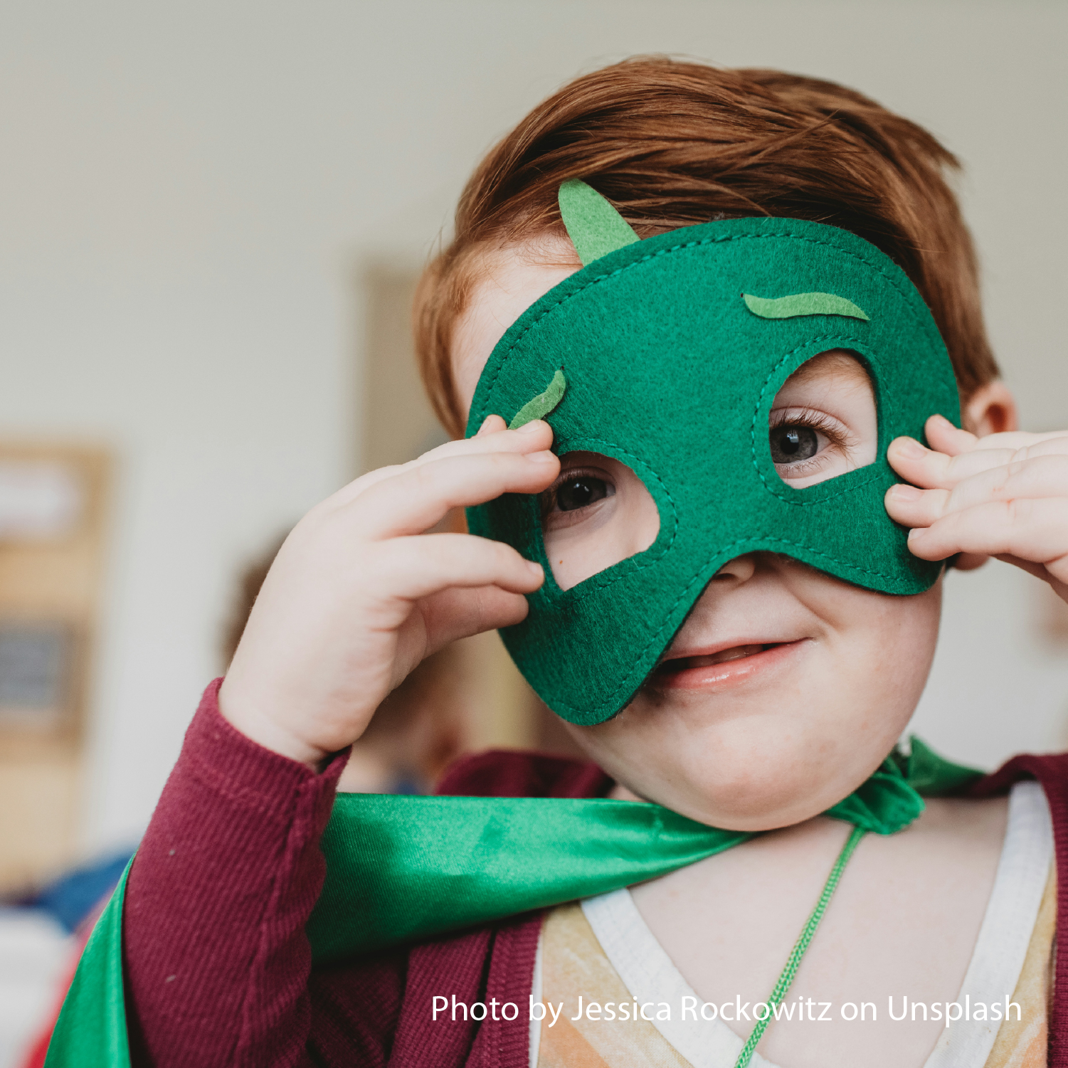 A child playfully wears a green superhero mask and cape, smiling with excitement. The setting is indoors, with a blurred background, creating a whimsical mood.