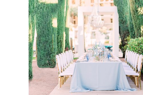 Elegant outdoor dining setup with a long table, white chairs, blue tablecloth, candles, and chandeliers amid tall green trees.