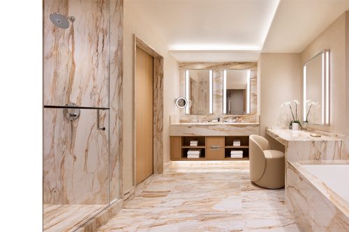 Luxurious marble bathroom with beige tones features a glass shower, large mirrors, vanity with towels, elegant lighting, and fresh white flowers.