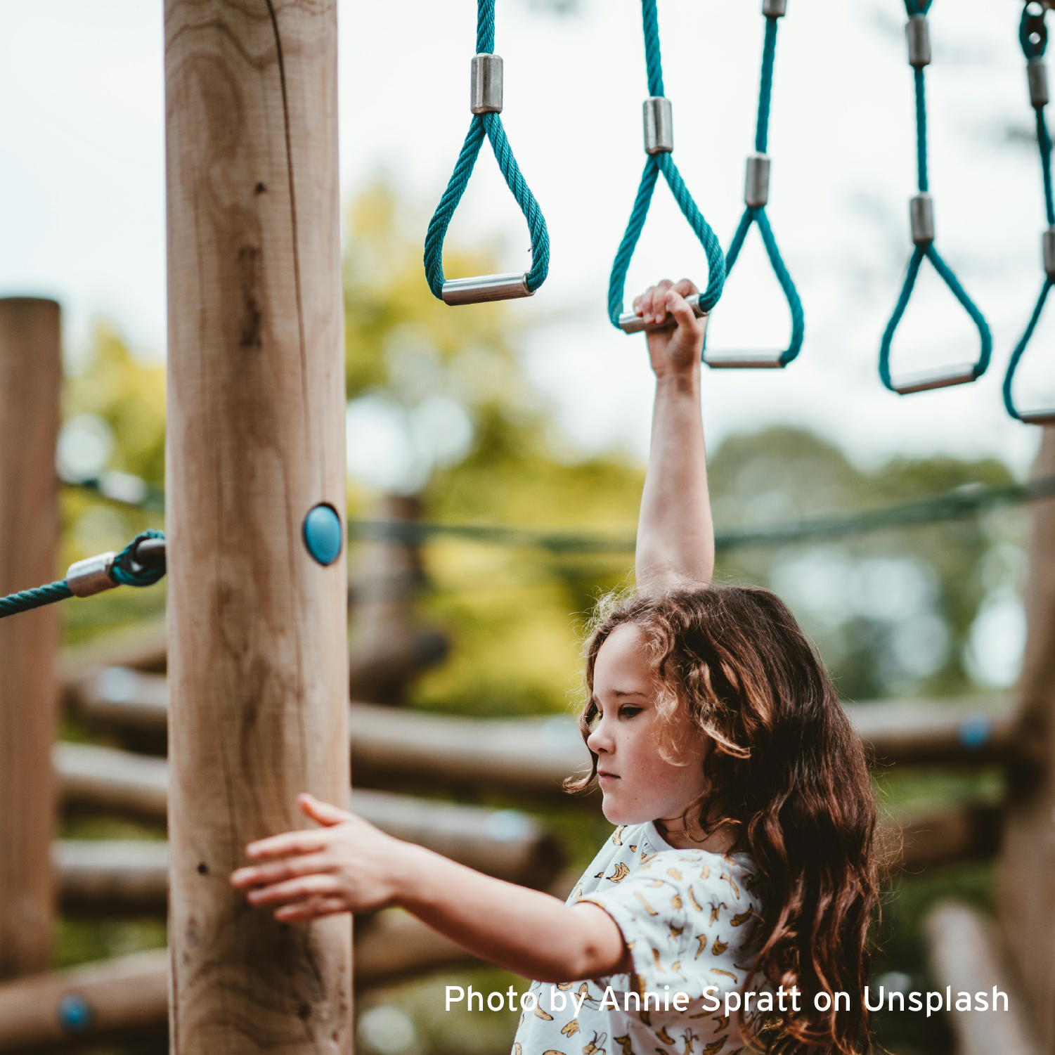 A young girl with curly hair confidently swings on blue monkey bars in a playground, demonstrating focus and determination against a blurred natural background.