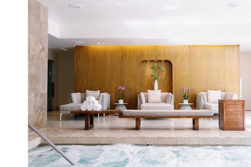 Modern spa lounge with three armchairs, rolled white towels, orchids, and a wooden wall feature. Foreground shows the edge of a bubbling spa pool.
